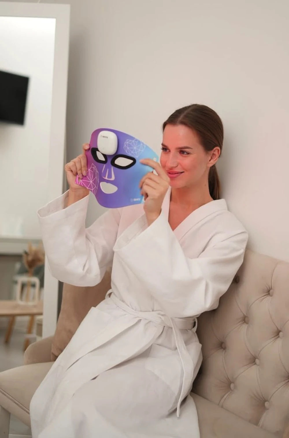 A woman in a white robe holding the colorful Zemits MaiSkin LED mask at home